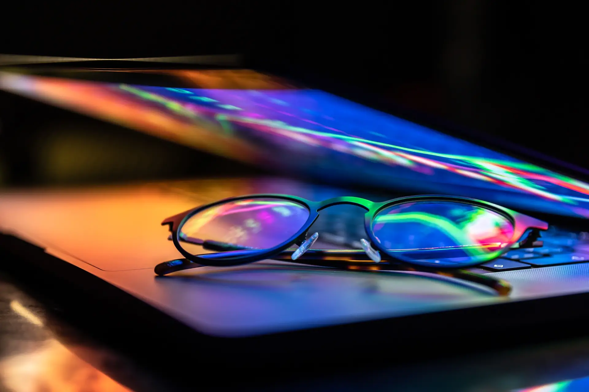 A laptop half closed in the dark with colourful glow and glasses.
